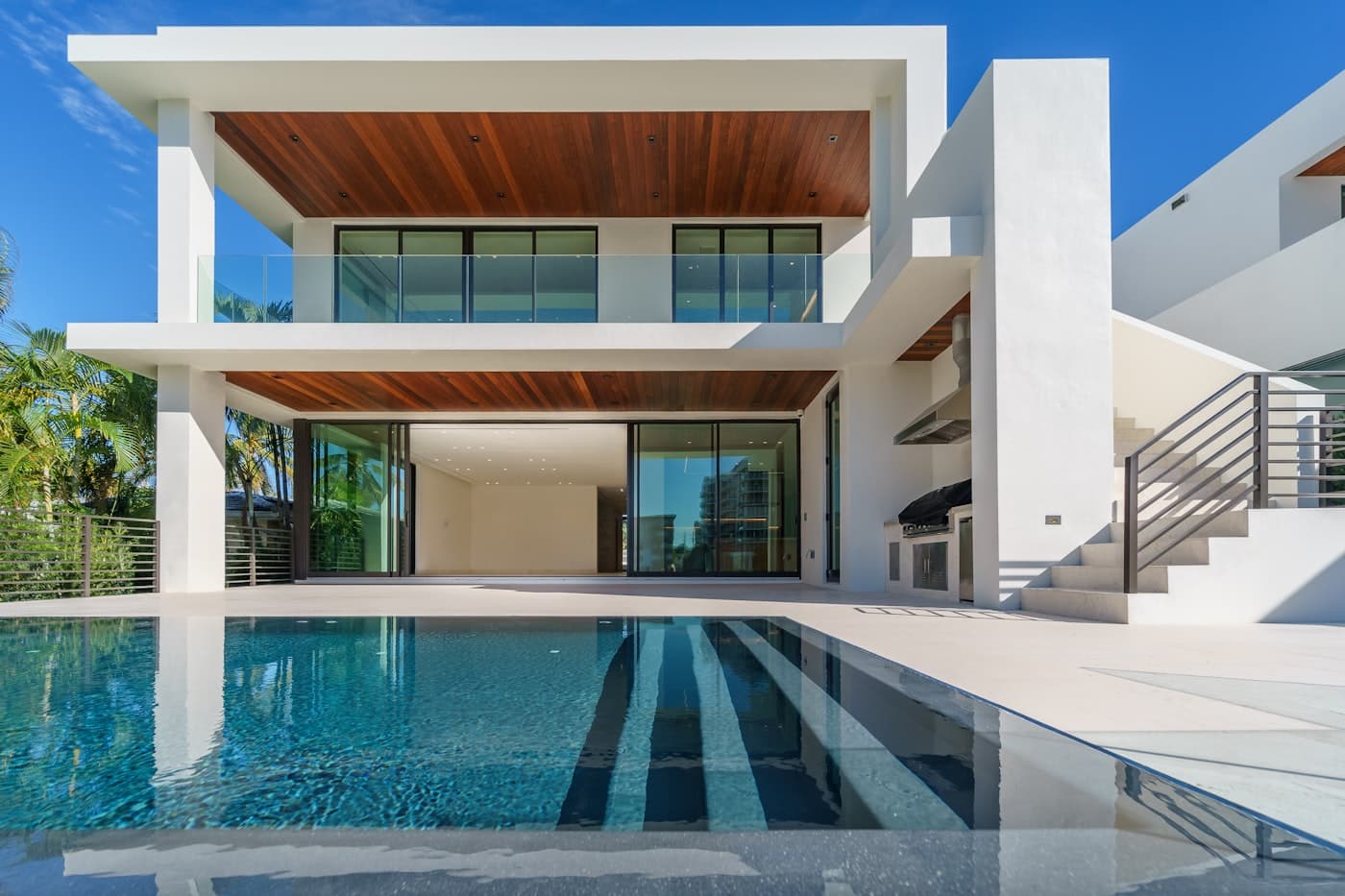 A beautiful modern home with a swimming pool alongside it, glass walls, and palm trees